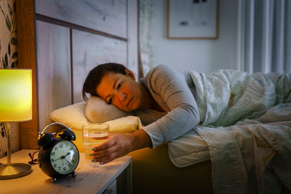 mature woman lying on bed during the night reaching a glass of water from night table selective focus on hand high resolution 42mp indoors digital capture taken with sony a7rii and zeiss batis 40mm f20 cf lens