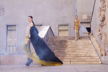 a model in a black cape, sheer taupe and yellow dress, and high boots walks in front of a marble staircase as another model in a sheer dress and thigh high boots descends