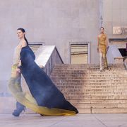a model in a black cape, sheer taupe and yellow dress, and high boots walks in front of a marble staircase as another model in a sheer dress and thigh high boots descends