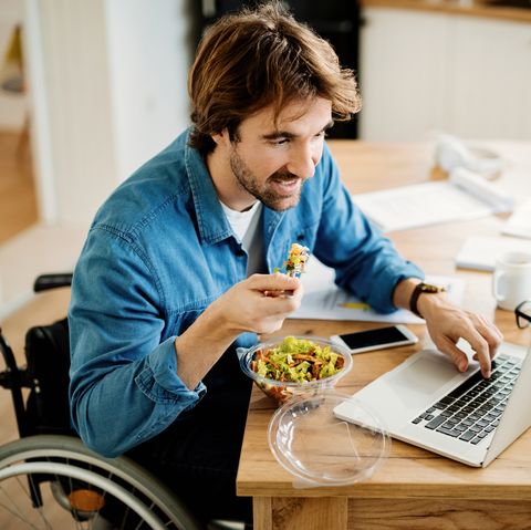 young businessman in wheelchair eating while surfing the net on laptop on lunch break at home