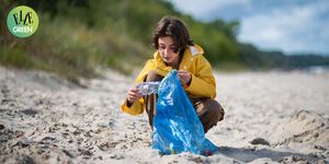 low angle view of adolescent girl in jacket picking up plastic garbage on sandy beach