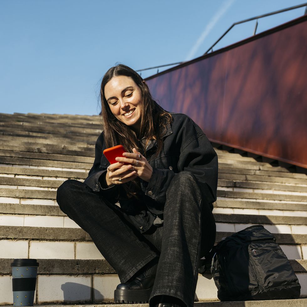 brunette young woman sitting on a public park stairs and texting on her red smartphone