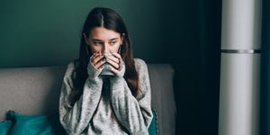 pensive, frost brunette woman in sweater, wrapped in green blue duvet sit on sofa and drink hot tea