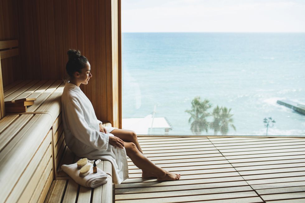 young beautiful slim woman in wooden dry sauna in spa centr in white bathrobe relaxation wellness and beauty concept finnish sauna for strengthening the bodys immune system, removing toxins, resting body and mind wooden eco sauna with a large window overlooking the ocean or sea
