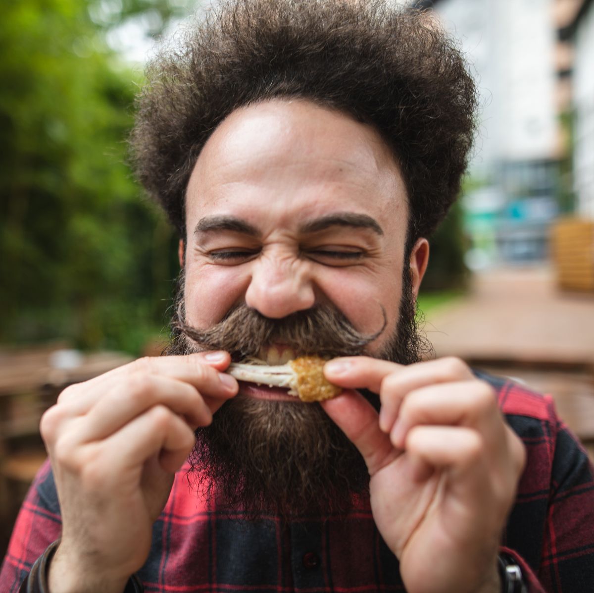 bearded hipster in a plaid shirt with pointy mustache eating a chicken wing