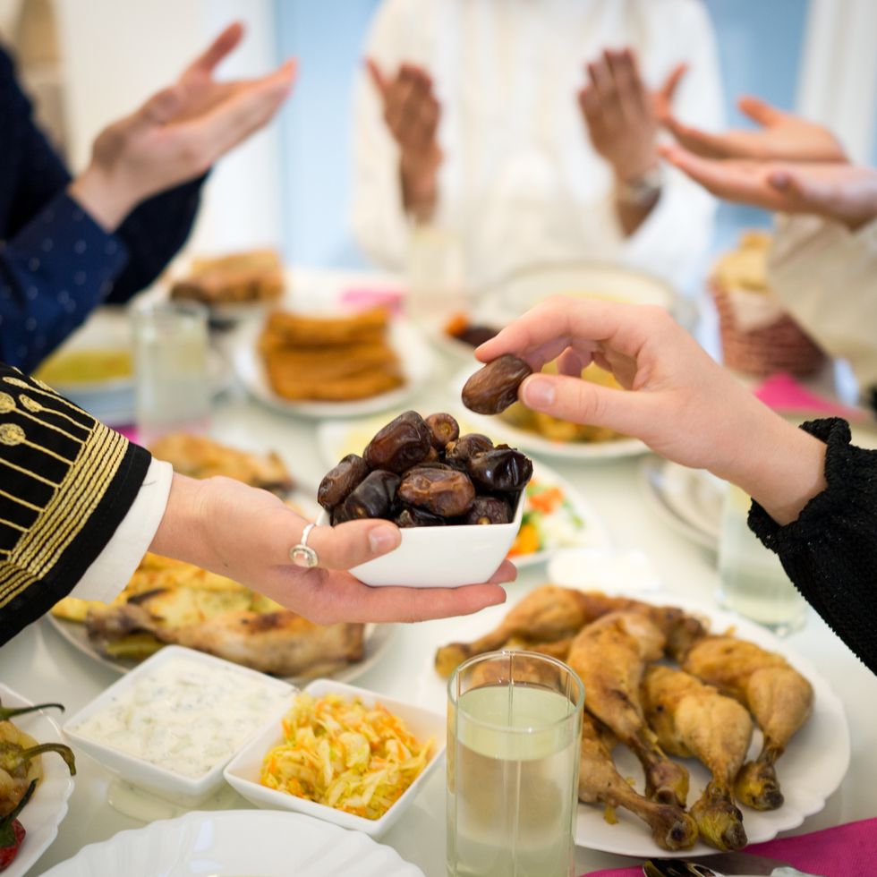 person holding out a bowl of dates for another person to reach for across a table with a feast laid out