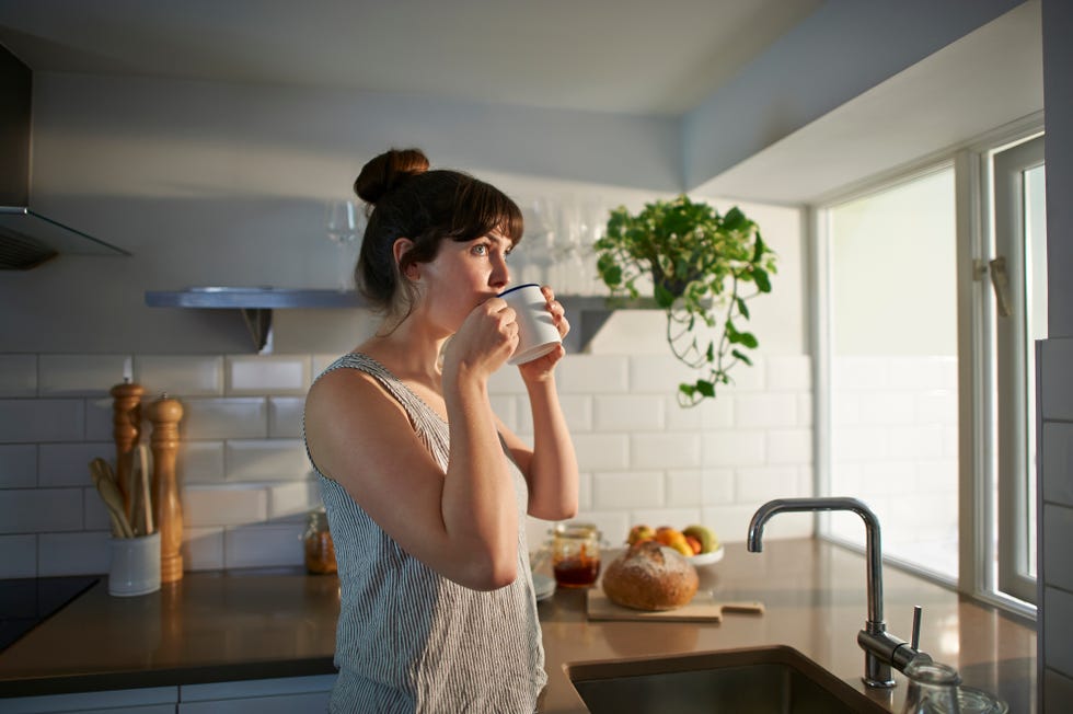 a woman standing in her kitchen drinks from a mug