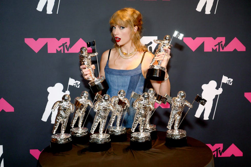 taylor swift holds two mtv video music awards as other statues sit on a table in front of her, she wears a sleeveless denim dress and several necklaces