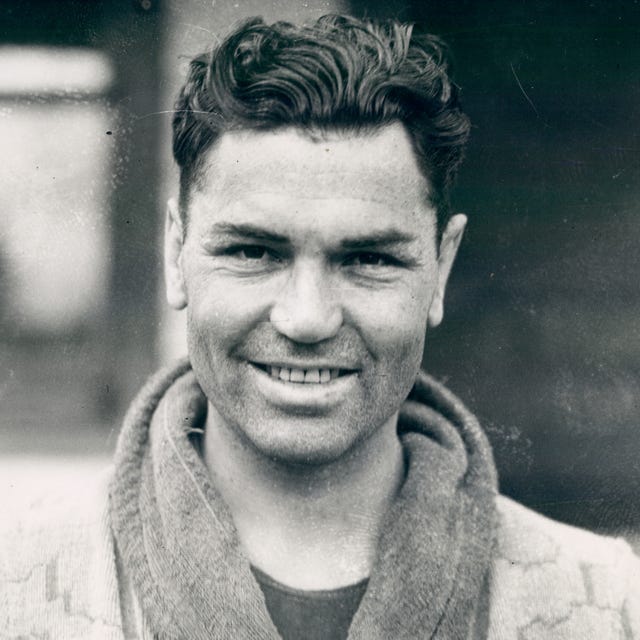 jack dempsey smiles at the camera, he wears a scarf, shirt and jacket