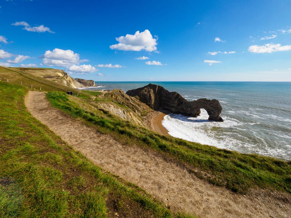 south west coast path at durdle door, dorset, uk. a grassy cliff path looking onto a small cove beach with a foamy tide coming in, a wall of rocks in the sea and blue skies.