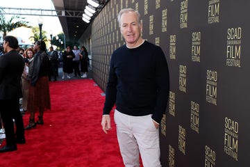 bob odenkirk smiles slightly at the camera, he wears a dark sweater and stands on a red carpet in front of a black backdrop