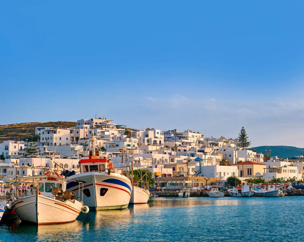 traditional greek fishing village and harbour on sunny morning or evening, boats moored by jetty and whitewashed houses alongside naoussa, paros island, greece clear blue sky, clouds, sunlight, colorful mediterranean landscape