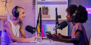 two interracial podcasters are sitting in a small broadcasting studio at home wearing headphones during the discussion and speaking at the microphones two fun girls recording a podcast
