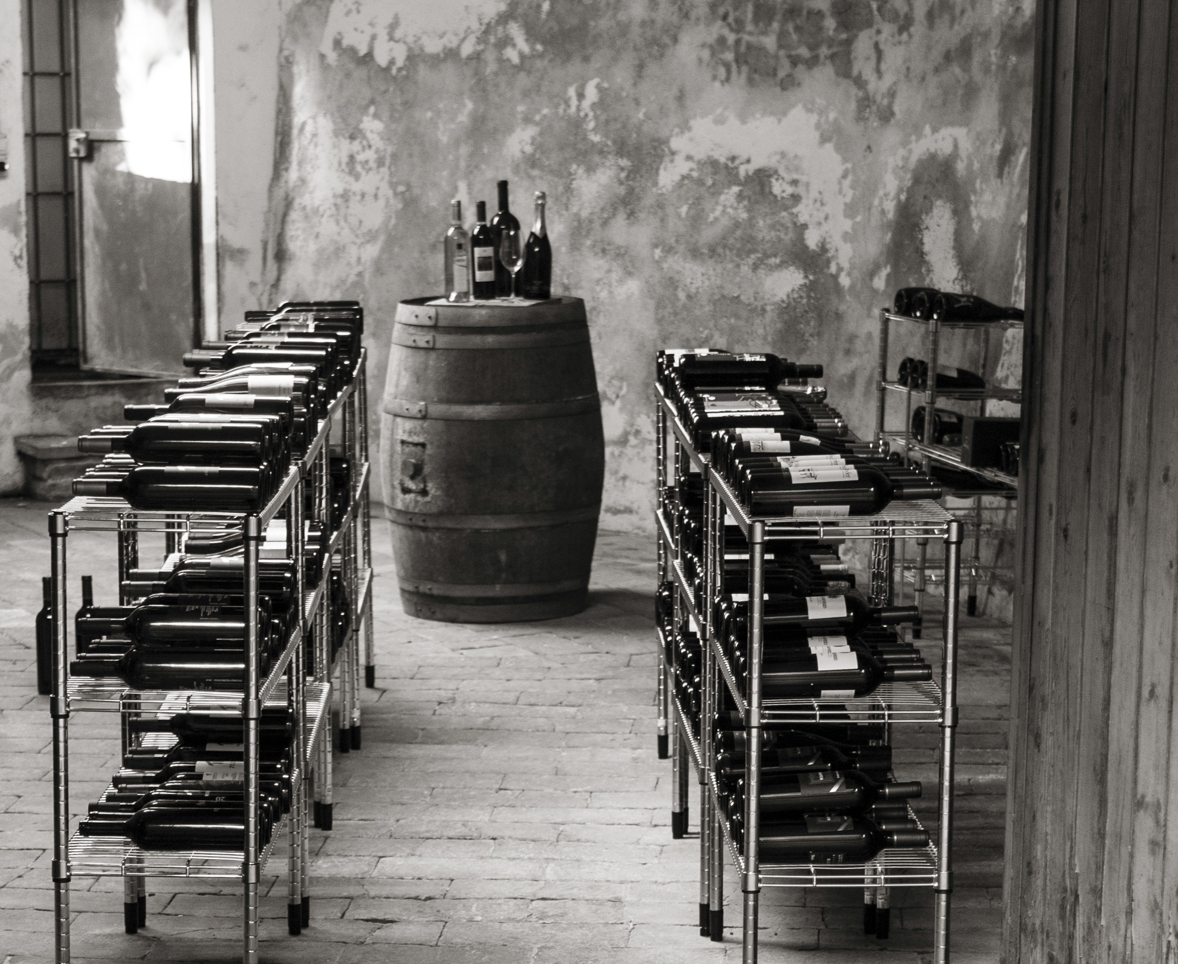 this is a black and white image of a wine cellar in a villa in the emilia romagna region of northern italy showing locally produced wines stored on stainless steel racks and a used wine cask used for tasting