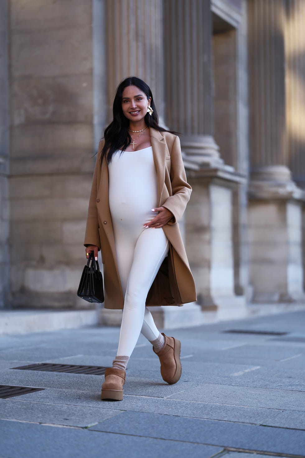 A pregnant woman walking in a stylish outfit with a coat and accessories