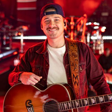 morgan wallen smiles while on a stage flooded with red light, he wears a backward baseball cap, jean jacket and white tshirt, a guitar hangs from a strap around his shoulders photo by christopher polkpenske media via getty images