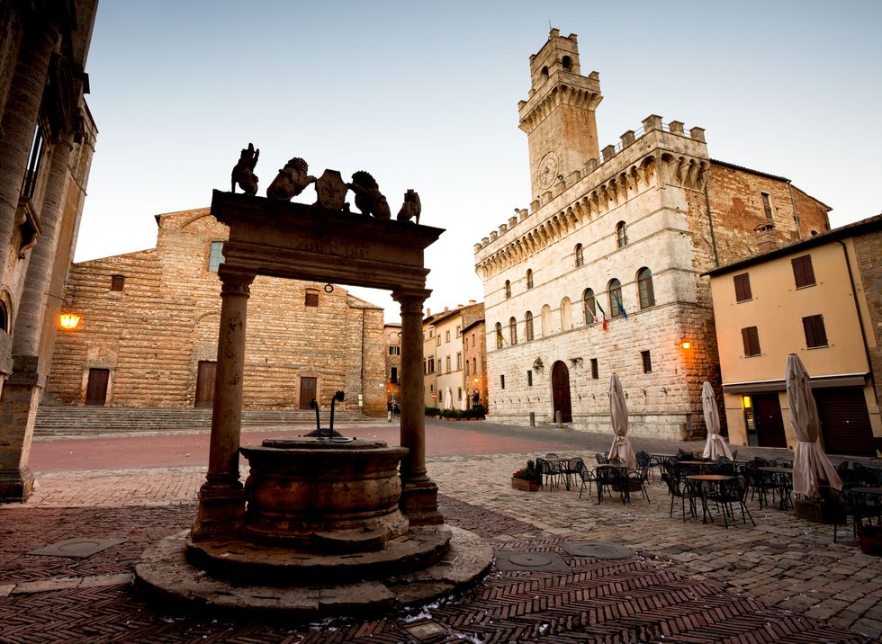 an ornate well silhouette stands in the foreground of the town plaza in montepulciano, italy in the background, the prominant town hall tower is starting to be illuminated with pre dawn light