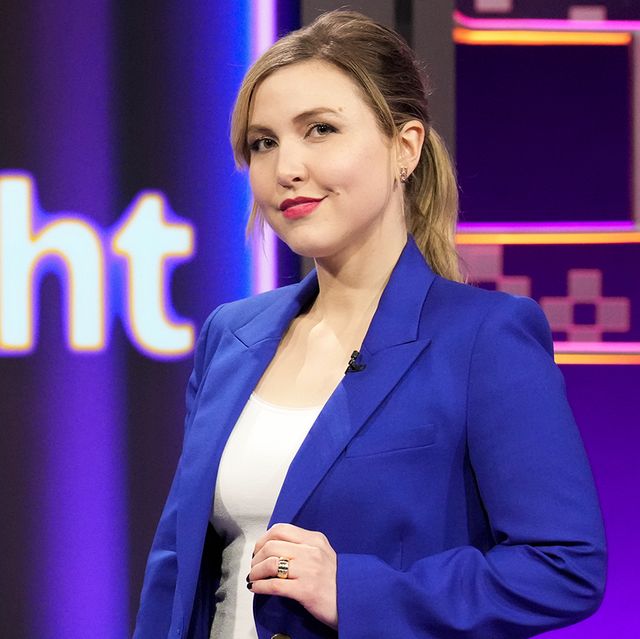 taylor tomlinson stands in front of a colorful background and smiles as she looks at the camera, she wears a royal blue suit jacket over a white top and holds one side of her jacket