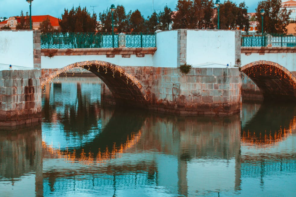 Il ponte romano di Tavira al tramonto, con luci decorative attorno agli archi del ponte. Il fiume Gilao scorre dolcemente, con solo qualche increspatura sulla superficie.