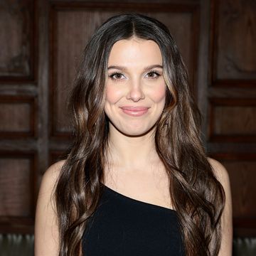 millie bobby brown smiles with her brown hair down and wearing a black one shoulder dress