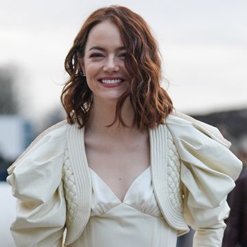 emma stone smiles at the camera as she stands outside, she wears a pale yellow full sleeved cropped jacket over a matching camisole