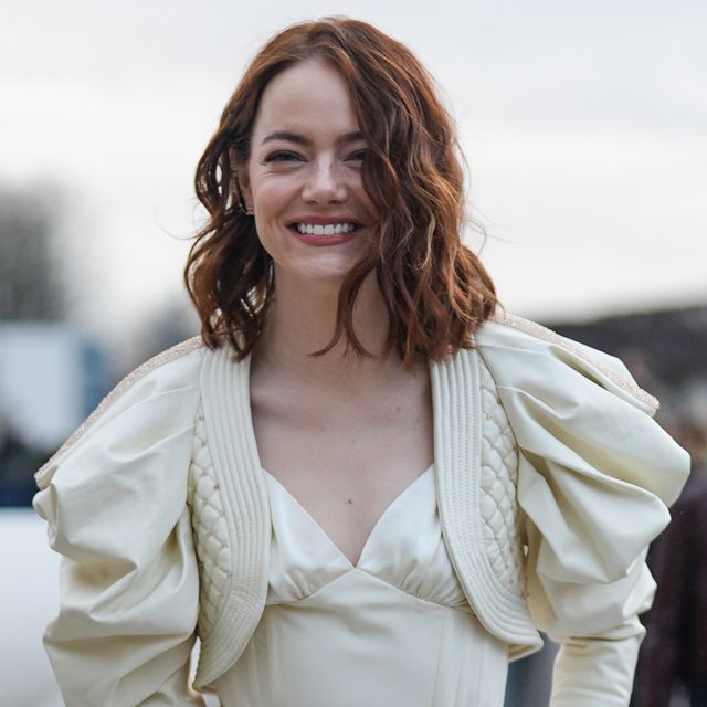 emma stone smiles at the camera as she stands outside, she wears a pale yellow full sleeved cropped jacket over a matching camisole