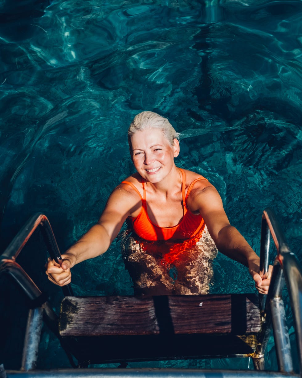 young stylish woman with short blonde hair in a bright orange swimsuit climbing a ladder out of clear blue water, smiling