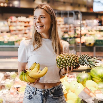 De voordelen van ananas een vrouw staat in een supermarkt met in haar linkerhand een ananas en in haar rechterhand een tros bananen, terwijl ze lachend naar rechts kijkt