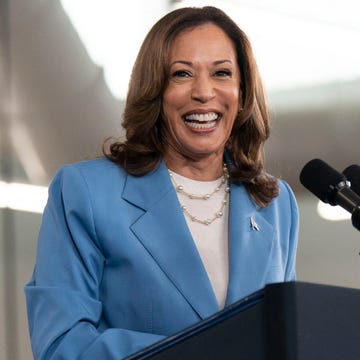 kamala harris smiles at the camera as she stands behind a podium with microphones, she wears a blue suit jacket, white blouse and pearl necklace