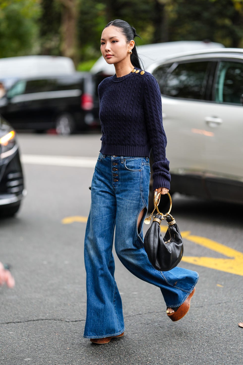 paris, france september 26 a guest wears gold earrings, navy blue knit sweater with gold detail, navy blue denim jean cutout pants, dark brown chloe platform shoes, shiny black chloe leather bag with gold chain detail, outside chloe, during the paris fashion week springsummer 2025 on september 26, 2024 in paris, france photo by edward berthelotgetty images