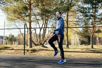 man in a blue hoodie and sportswear practices high knee exercises on a clear day in a public park. his active lifestyle is highlighted by the vibrant outdoor setting and focused energy.