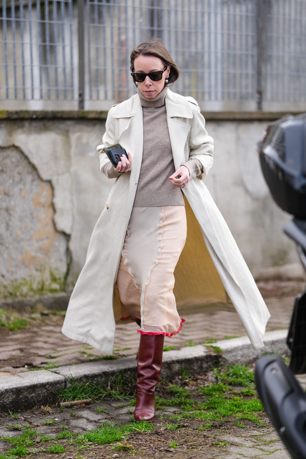 milan, italy february 26: a guest wears black ray ban sunglasses, silver earrings, light brown turtleneck long sleeve sweater, cream trench coat, mixed metal rings, light gray light pink sheer mesh midi skirt, shiny burgundy bordeaux slouchy knee high heeled boots leather shoes, outside jil sander, during the milan fashion week womens fall/winter 2025 2026 on february 26, 2025 in milan, italy. (photo by edward berthelot/getty images)