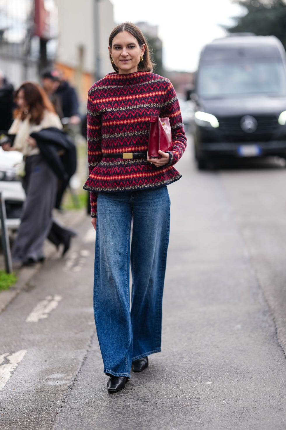 milan, italy march 01: a guest wears gold earrings, dark burgundy bordeaux pattern tailored sweater, shiny black leather belt, shiny burgundy bordeaux clutch leather bag, navy blue washed denim jean loose pants, shiny black boots leather shoes, outside ferragamo, during the milan fashion week womens fall/winter 2025 2026 on march 1, 2025 in milan, italy. (photo by edward berthelot/getty images)