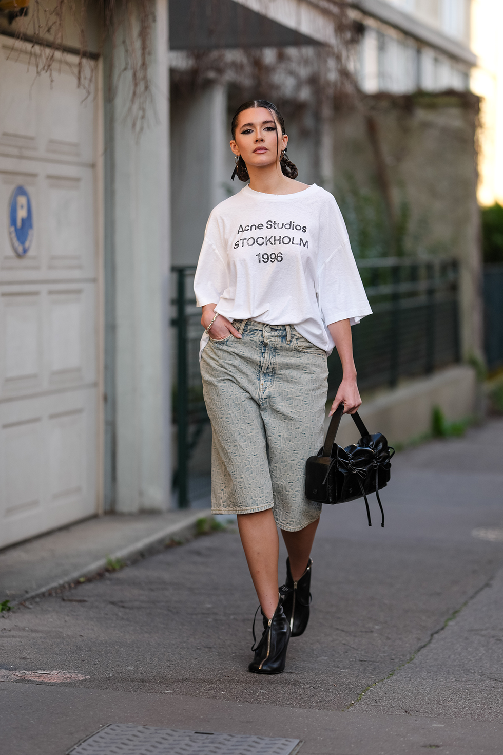 paris, france march 05: chiara king wears slingback braided pigtails, silver earrings, white oversized acne studios shirt, navy blue light brown washed denim jean jorts, a silver bracelet, shiny black acne studios leather bag, shiny black zipped heels leather shoes, outside acne studios, during the paris fashion week women's fall/winter 2025 2026 on march 5, 2025 in paris, france. (photo by edward berthelot/getty images)