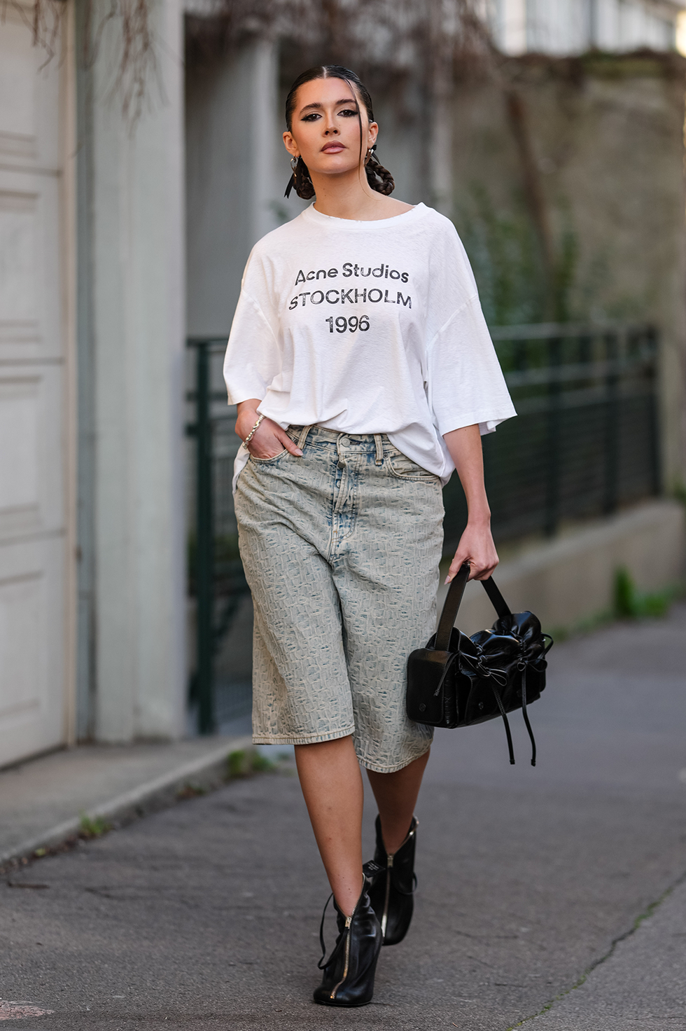 paris, france march 05: chiara king wears slingback braided pigtails, silver earrings, white oversized acne studios shirt, navy blue light brown washed denim jean jorts, a silver bracelet, shiny black acne studios leather bag, shiny black zipped heels leather shoes, outside acne studios, during the paris fashion week women's fall/winter 2025 2026 on march 5, 2025 in paris, france. (photo by edward berthelot/getty images)