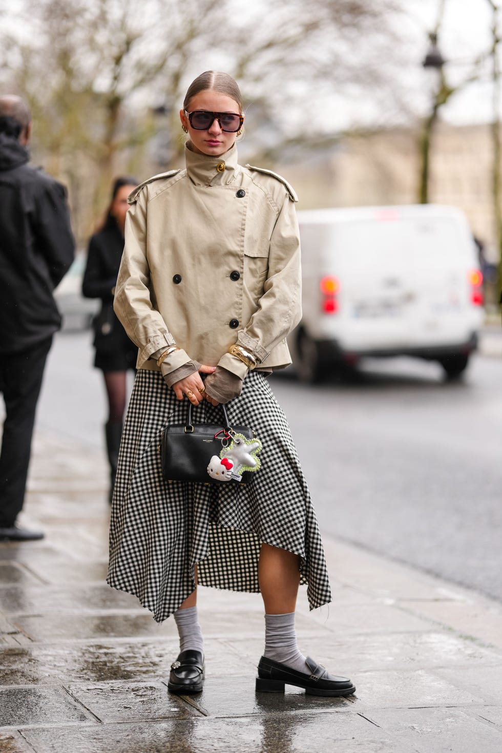 paris, france march 10: a guest wears dark brown sunglasses, gold earrings, light brown high neck double breasted trench jacket, gold bracelets, gold rings, white black pattern/print midi skirt, shiny black saint laurent leather bag, hello kitty bag charm, light gray socks, shiny black loafer moccasin leather shoes, silver metallic star bag charm, outside marine serre, during the paris fashion week womens fall/winter 2025 2026 on march 10, 2025 in paris, france. (photo by edward berthelot/getty images)
