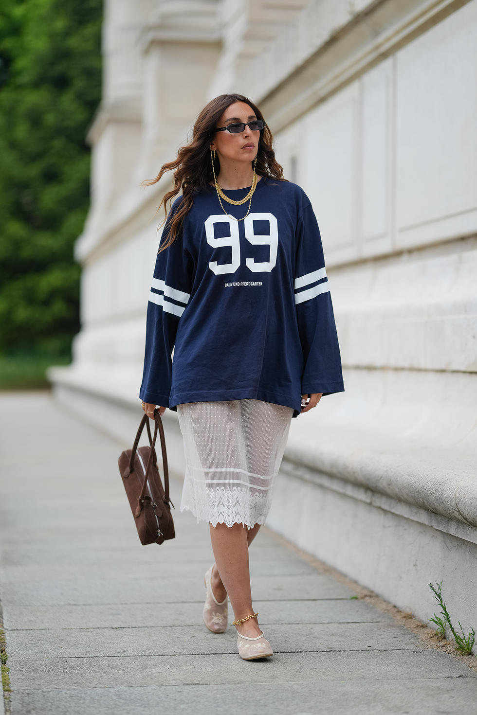 paris, france april 25: gabriella berdugo wears long, wavy brown hair with a center part, paired with a matte complexion, nude lipstick and natural brows. black rectangular sunglasses are styled with layered gold necklaces. a navy oversized long sleeve t shirt with white stripes and the number "99" printed on the front by baum is worn over a semi sheer white lace midi skirt, also by baum. a brown suede baguette style handbag by alaia is carried in hand. beige mesh ballet flats by anthropologie are worn with a thin gold anklet. full body shot, during a street style fashion photo session, on april 25, 2025 in paris, france. (photo by edward berthelot/getty images)