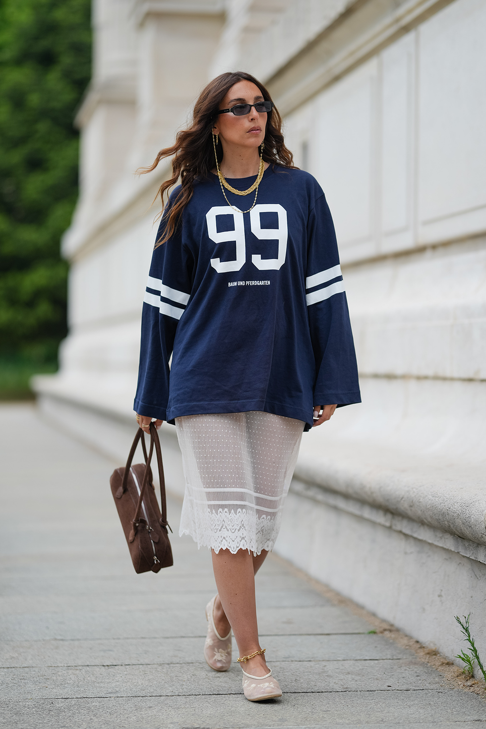 paris, france april 25: gabriella berdugo wears long, wavy brown hair with a center part, paired with a matte complexion, nude lipstick and natural brows. black rectangular sunglasses are styled with layered gold necklaces. a navy oversized long sleeve t shirt with white stripes and the number "99" printed on the front by baum is worn over a semi sheer white lace midi skirt, also by baum. a brown suede baguette style handbag by alaia is carried in hand. beige mesh ballet flats by anthropologie are worn with a thin gold anklet. full body shot, during a street style fashion photo session, on april 25, 2025 in paris, france. (photo by edward berthelot/getty images)