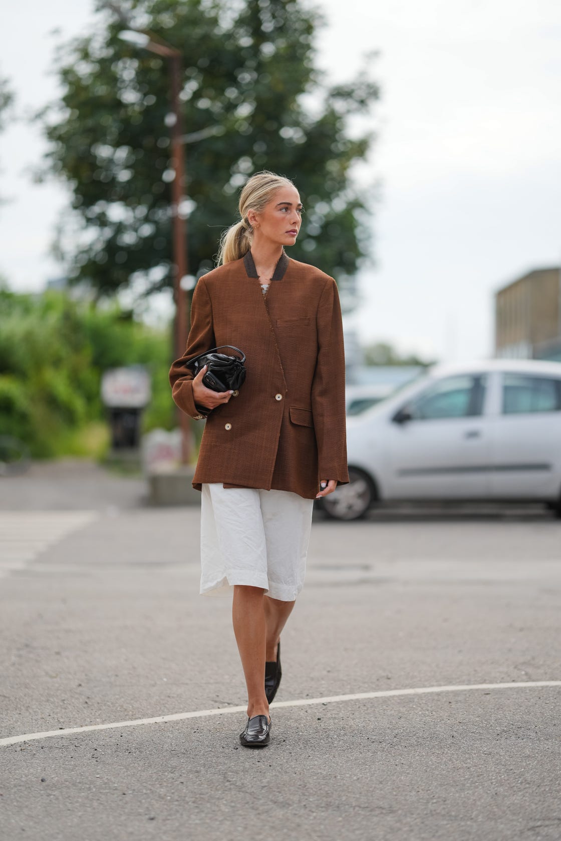 copenhagen, denmark august 07: a guest wears light blonde hair styled in a sleek low ponytail with a center part, an oversized mid brown textured blazer jacket with a contrasting dark brown knit collar, off center button fastening with light buttons, a left chest welt pocket and a right flap pocket, a white layer peeking at the neckline, loose white knee length bermuda shorts in crisp cotton, and black polished leather loafers with low stacked heels and rounded toes, while a small black leather ruched pouch clutch with a short handle is carried in one hand, walking, outside marimekko, during copenhagen fashion week, on august 07, 2025 in copenhagen, denmark (photo by edward berthelot/getty images)