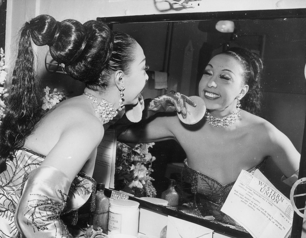 American-born cabaret singer Josephine Baker (1906 - 1975) applies makeup with a powder puff in her dressing room before a performance at the Strand Theatre. A Western Union telegram from jazz singer Sophie Tucker is attached to the mirror. Baker has her hair pulled back into a fall and wears an evening dress, matching long satin gloves and a jeweled choker.