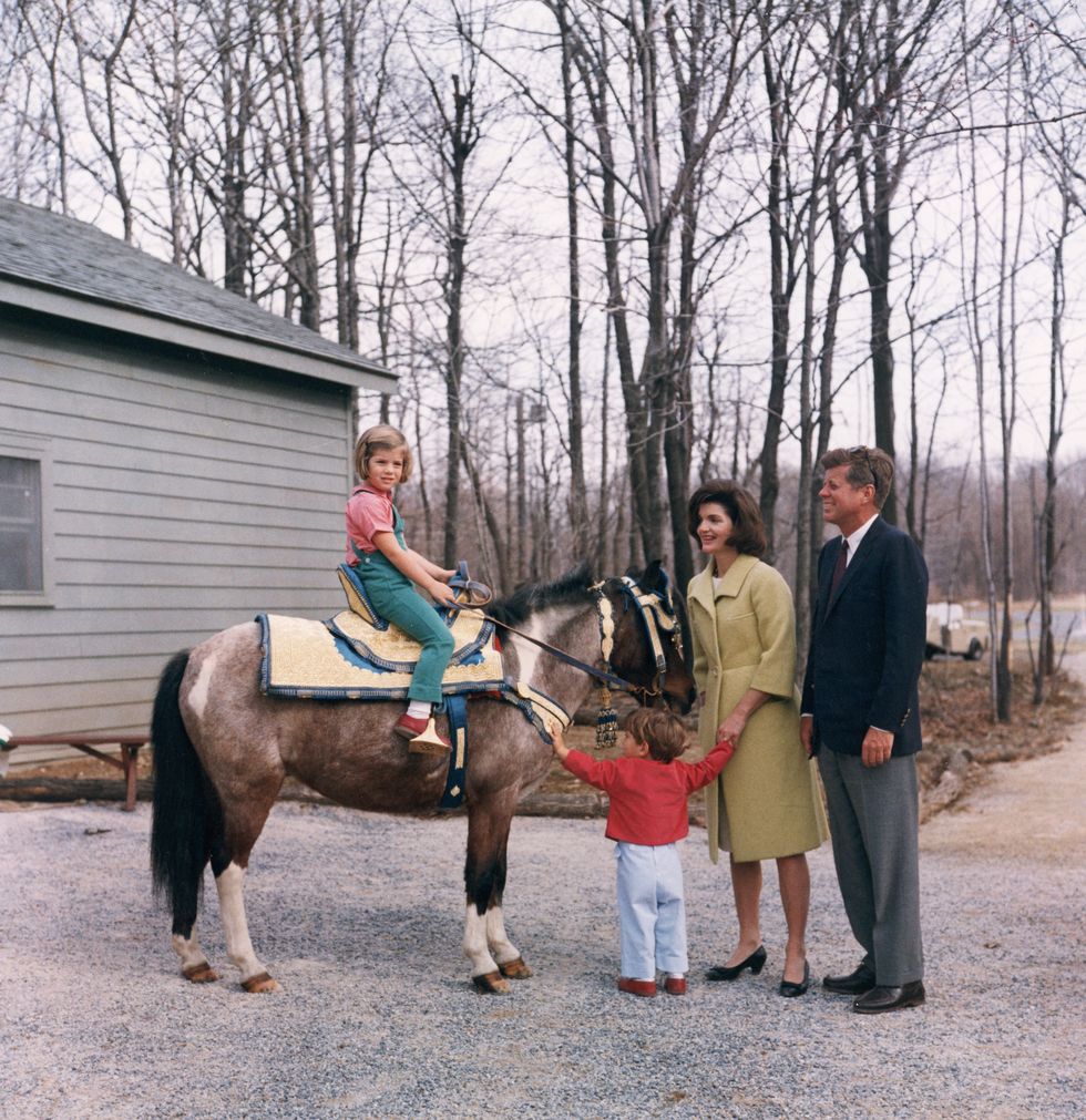 John F Kennedy stands outdoors with his wife, Jacqueline Kennedy, as their daughter, Caroline, sits atop a pony and their son, John Jr., holds his arms out.