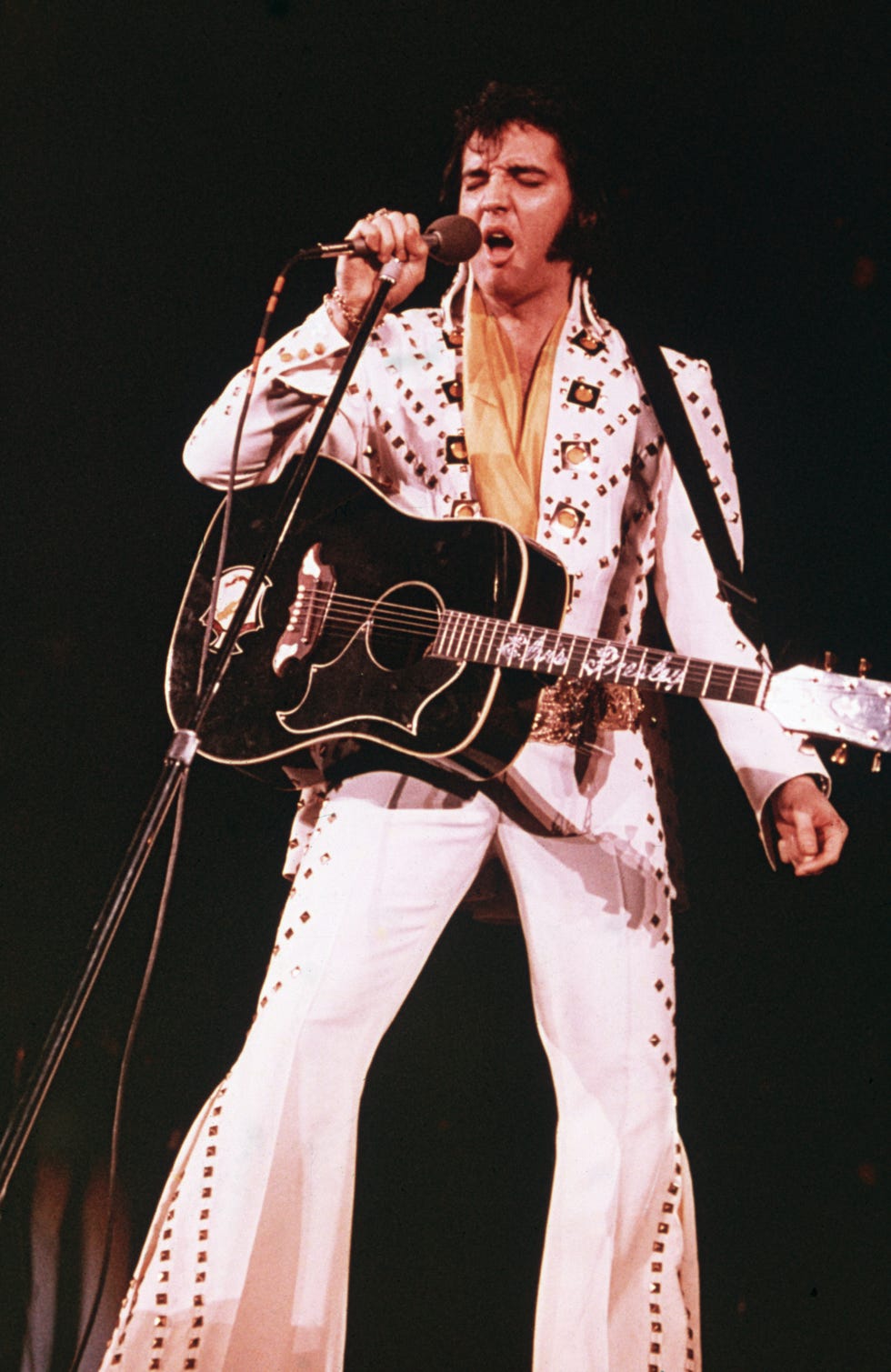 Circa 1975 American rock singer Elvis Presley 1935 1977, wearing a white rhinestone studded suit and strappy guitar, singing into a microphone with his eyes closed Photo International via Getty Images