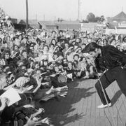 Elvis Presley performs outdoors on a small stage to the adulation of a young crowd.