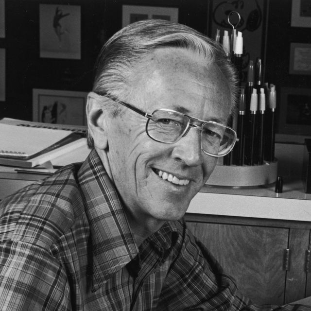 charles schulz smiling at the camera while seated, he wears wire rimmed large glasses and a plaid collared shirt, behind him are drawing supplies and framed artwork on a wall