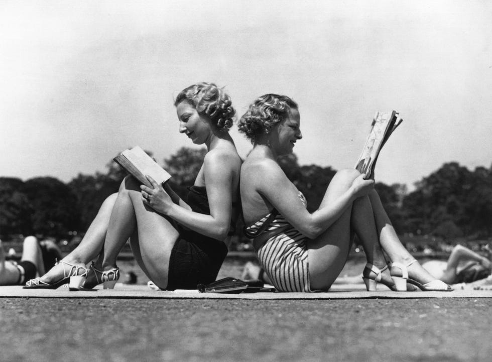 two london women reading back to back as they sunbathe by the banks of the serpentine in hyde park, london