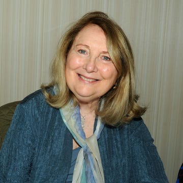 teri garr smiles at the camera, she wears a blue sweater and tank top with a multicolor scarf and necklace