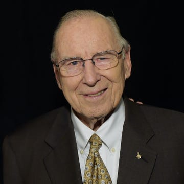 james a lovell jr smiles at the camera, he wears glasses, a white collared shirt, black suit jacket and yellow patterned tie