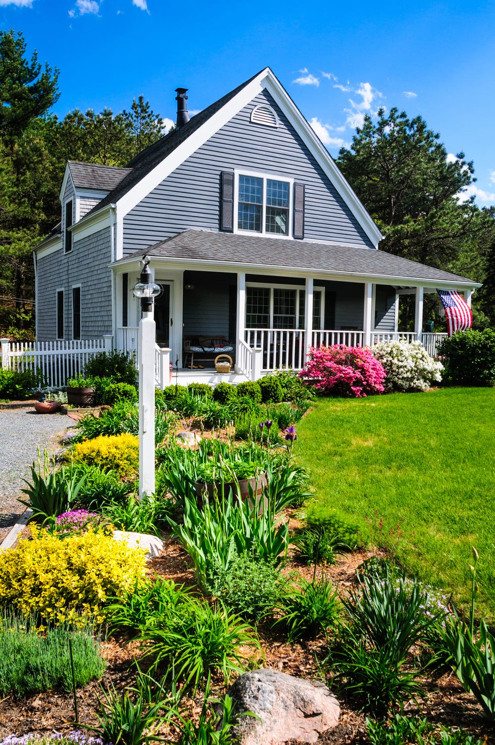 an american flag flies from the open porch and gardens surround a small  single family home on a spring afternoon on cape cod on the massachusetts coast property release attached