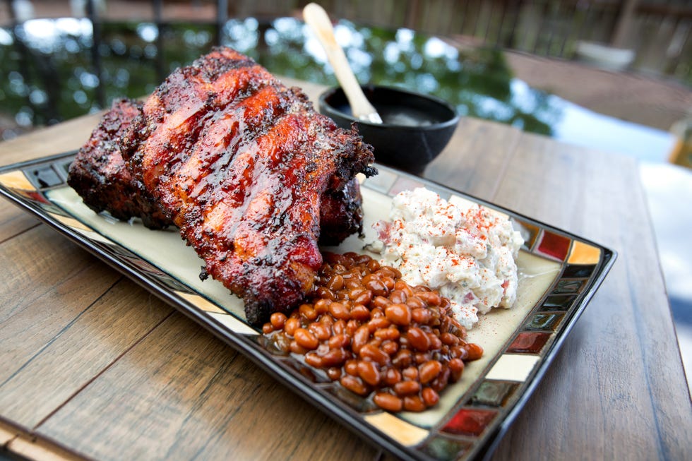 platter with a full rack of stacked baby back ribs with potato salad and baked beans on the side plate is on a table outside in a backyard