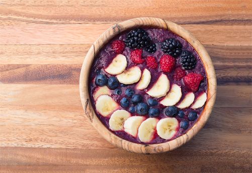 berry acai smoothie bowl topped with bananas, blueberries raspberries and blackberries in a wooden bowl
